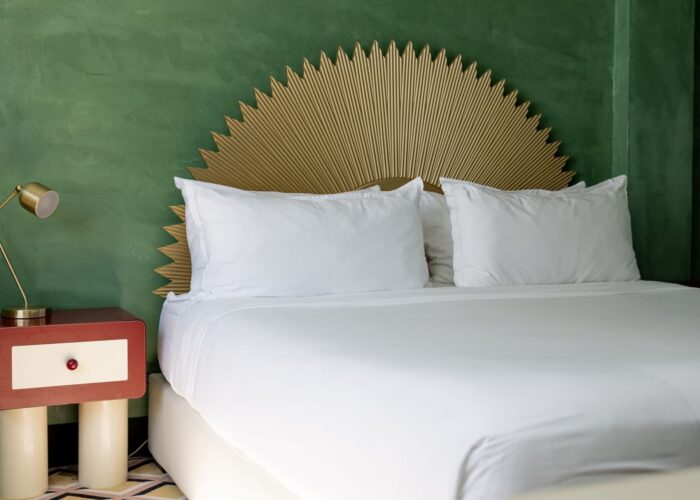 Bed under a gold sunburst headboard by a red nightstand with a lamp in the Eclectic King Room at Crest Hotel & Suites