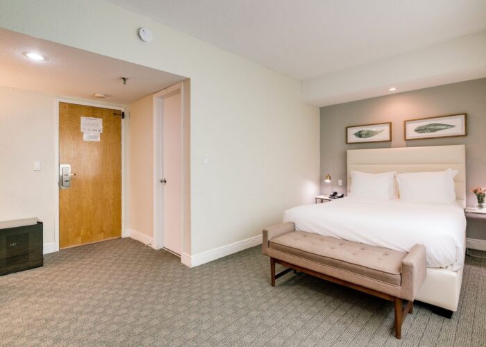 Bed under framed art prints with a tan bench by a wooden entryway door in the Standard Queen Room at Crest Hotel & Suite